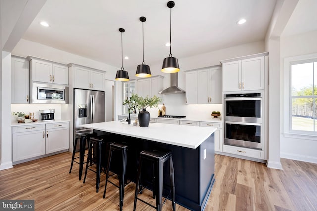 kitchen featuring wall chimney range hood, pendant lighting, a center island with sink, and white cabinets