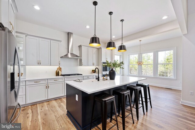 kitchen with appliances with stainless steel finishes, light hardwood / wood-style flooring, wall chimney exhaust hood, an island with sink, and pendant lighting