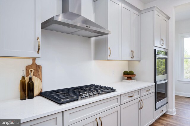 kitchen featuring light wood-type flooring, gas stovetop, stainless steel double oven, and wall chimney exhaust hood