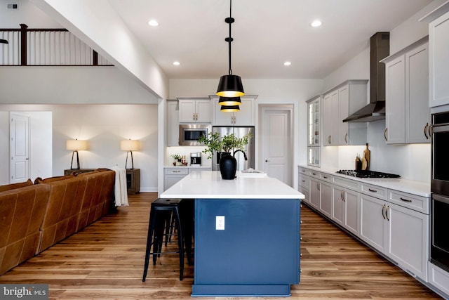 kitchen featuring a kitchen breakfast bar, light wood-type flooring, appliances with stainless steel finishes, a center island with sink, and wall chimney range hood