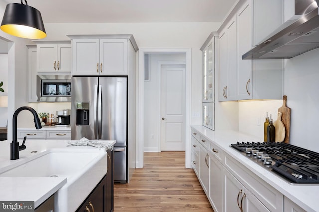 kitchen with wall chimney exhaust hood, hanging light fixtures, white cabinetry, appliances with stainless steel finishes, and light hardwood / wood-style floors