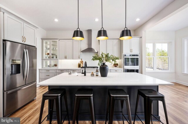 kitchen featuring a kitchen breakfast bar, light wood-type flooring, appliances with stainless steel finishes, an island with sink, and wall chimney range hood