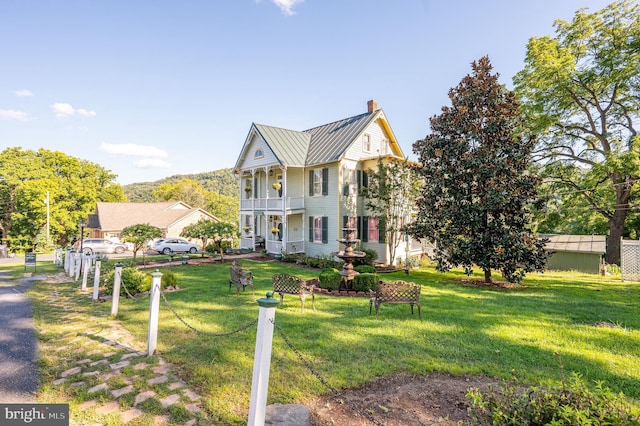 view of front of home with a balcony, a chimney, metal roof, a standing seam roof, and a front lawn