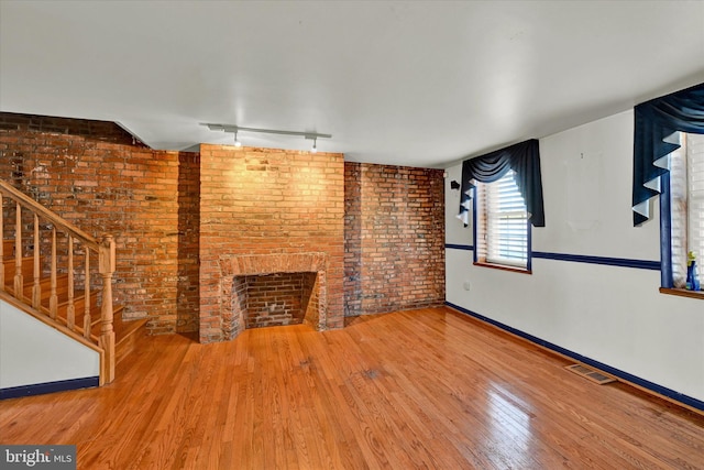 unfurnished living room featuring hardwood / wood-style flooring, brick wall, track lighting, and a brick fireplace