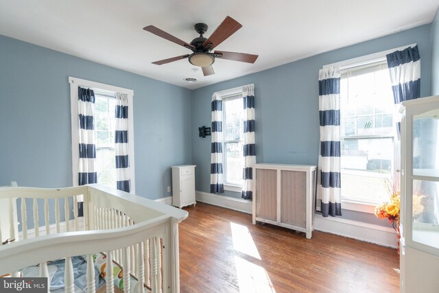 bedroom featuring ceiling fan, wood-type flooring, a nursery area, and multiple windows