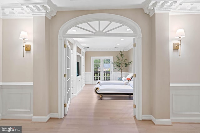 corridor with light wood finished floors, a wainscoted wall, crown molding, french doors, and a decorative wall