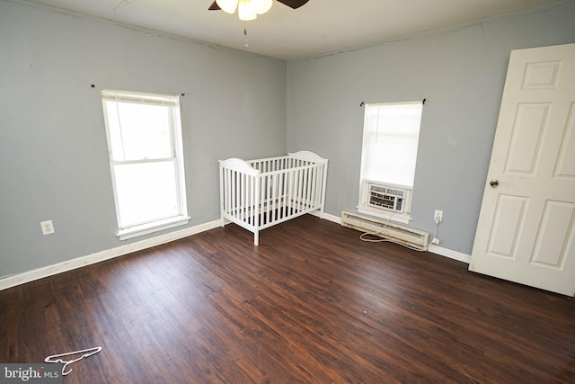 unfurnished bedroom with ceiling fan, baseboard heating, a nursery area, and hardwood / wood-style flooring