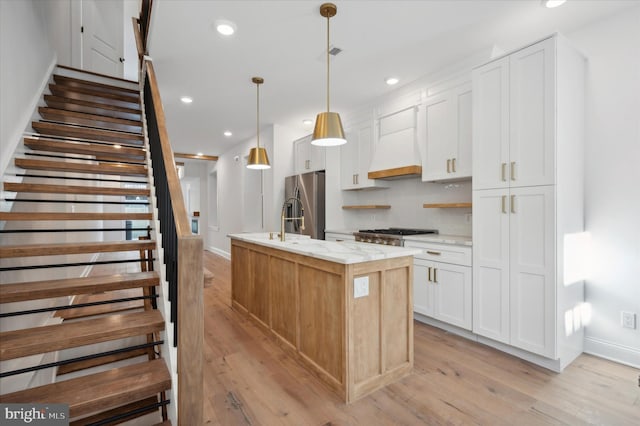 kitchen featuring custom range hood, a kitchen island with sink, decorative light fixtures, light hardwood / wood-style flooring, and white cabinets