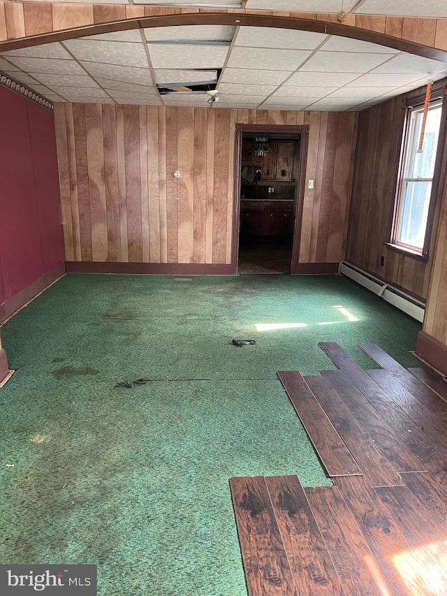 carpeted empty room featuring a paneled ceiling, wooden walls, and baseboard heating