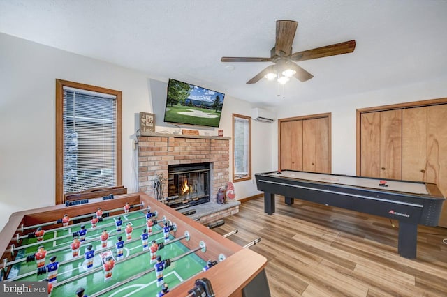 recreation room with a brick fireplace, ceiling fan, light wood-type flooring, and a wall unit AC