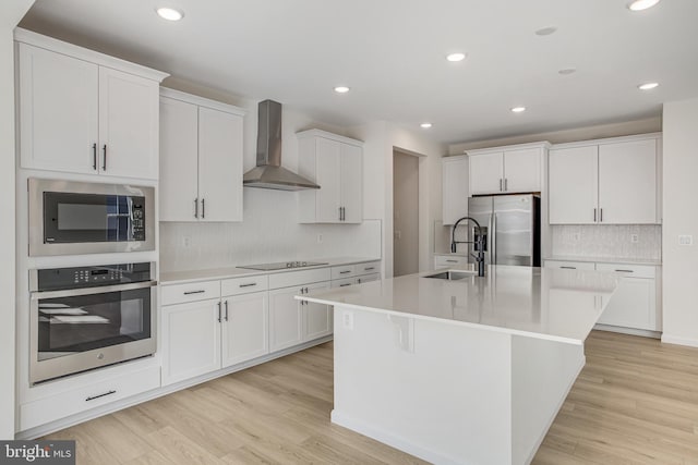 kitchen with white cabinets, wall chimney exhaust hood, stainless steel appliances, light wood-style floors, and a sink