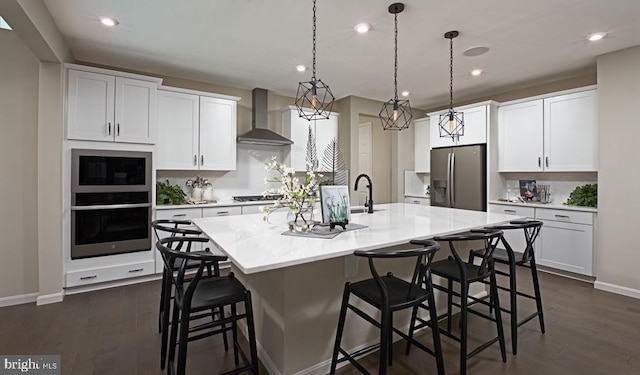 kitchen with appliances with stainless steel finishes, wall chimney range hood, an island with sink, and dark wood-style floors