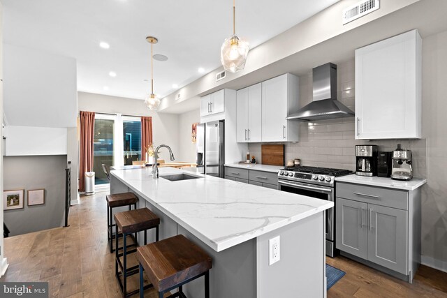 kitchen featuring wall chimney range hood, a kitchen island with sink, gray cabinetry, sink, and appliances with stainless steel finishes