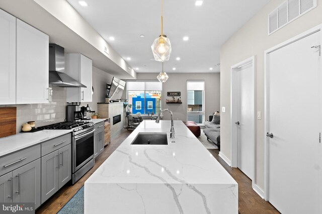 kitchen featuring pendant lighting, wall chimney range hood, an island with sink, and stainless steel gas range oven