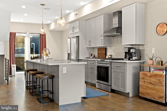 kitchen with wall chimney range hood, sink, dark hardwood / wood-style floors, appliances with stainless steel finishes, and an island with sink