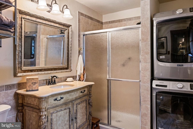 bathroom featuring stacked washer / drying machine, a shower with shower door, vanity, tile walls, and toilet