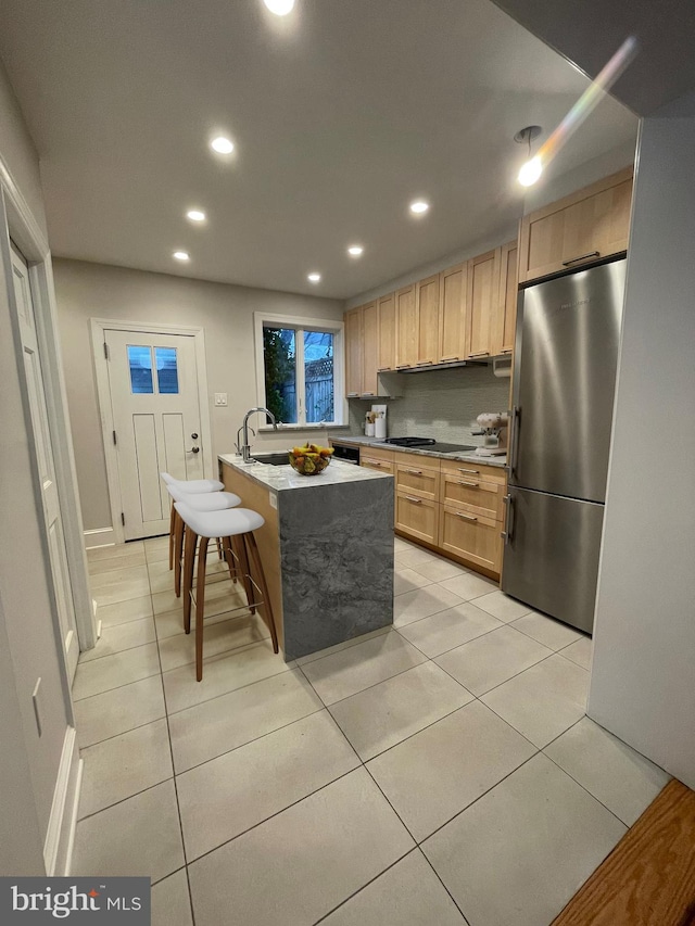 kitchen with light brown cabinets, stainless steel fridge, an island with sink, light tile patterned flooring, and a kitchen bar