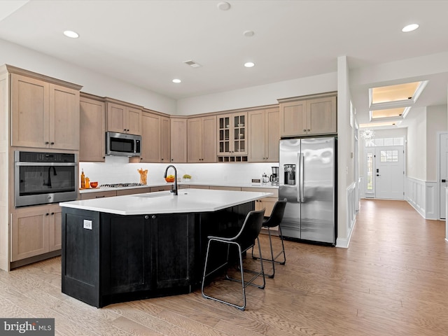 kitchen featuring light wood-type flooring, a kitchen island with sink, backsplash, sink, and appliances with stainless steel finishes