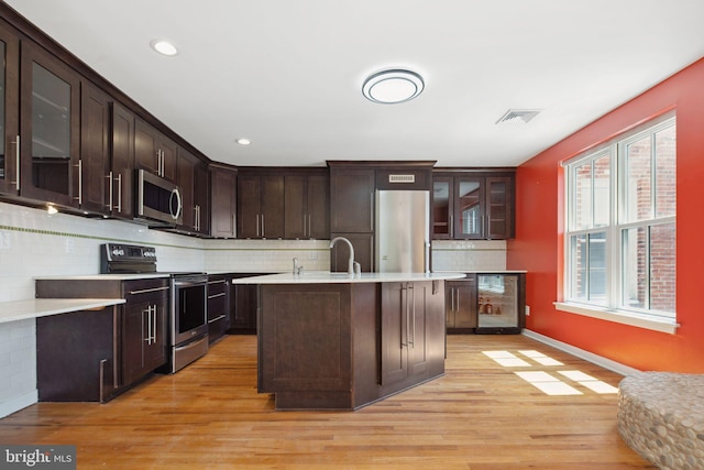 kitchen with backsplash, stainless steel appliances, light hardwood / wood-style flooring, dark brown cabinets, and beverage cooler