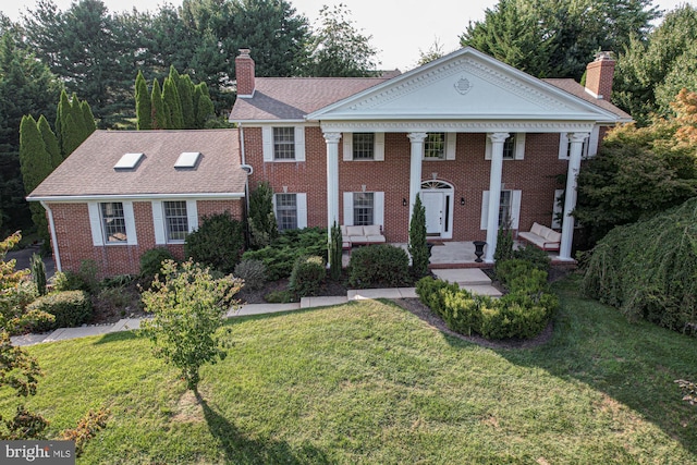 greek revival inspired property featuring covered porch, brick siding, roof with shingles, a chimney, and a front yard