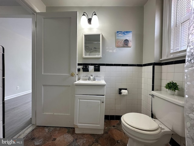 bathroom with toilet, a wainscoted wall, stone finish floor, vanity, and tile walls