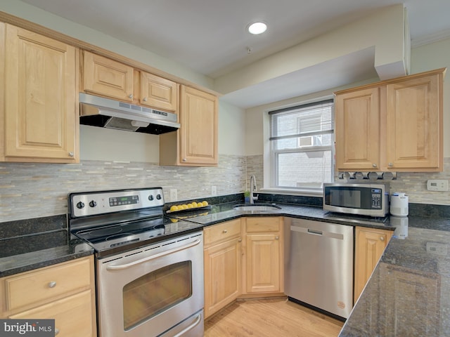 kitchen with light brown cabinets, under cabinet range hood, a sink, appliances with stainless steel finishes, and dark stone countertops