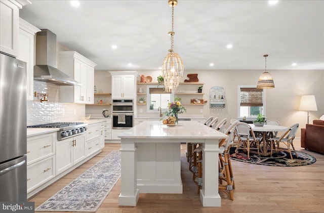 kitchen with wall chimney range hood, hanging light fixtures, appliances with stainless steel finishes, a breakfast bar area, and light hardwood / wood-style floors