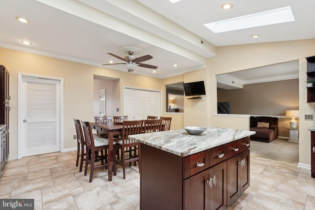 kitchen with ceiling fan, lofted ceiling with skylight, a kitchen island, dark brown cabinets, and light stone countertops