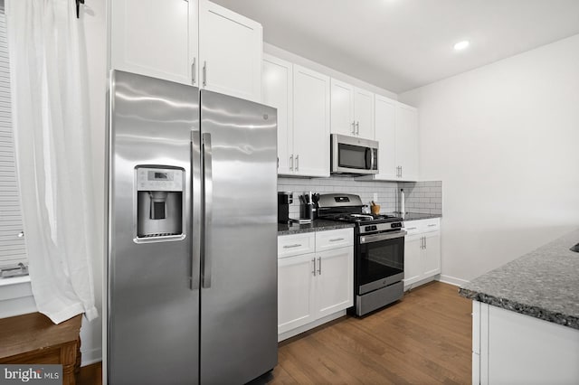 kitchen with tasteful backsplash, wood-type flooring, white cabinets, appliances with stainless steel finishes, and dark stone countertops