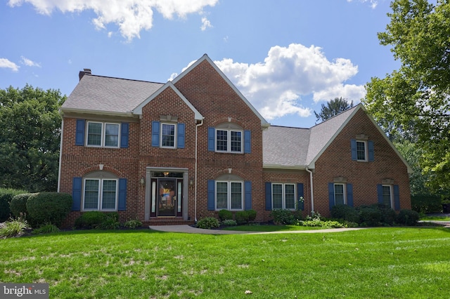 colonial house featuring a front yard, brick siding, roof with shingles, and a chimney