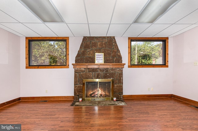 unfurnished living room with a stone fireplace, a drop ceiling, wood finished floors, and baseboards