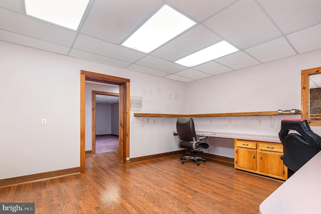 unfurnished office with baseboards, built in study area, a drop ceiling, and wood finished floors