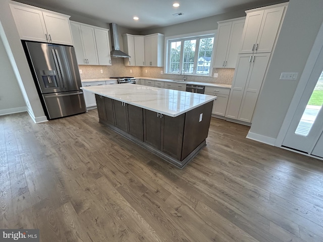 kitchen with appliances with stainless steel finishes, white cabinetry, a kitchen island, hardwood / wood-style flooring, and wall chimney range hood