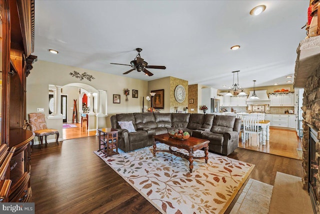 living room featuring ceiling fan, dark wood-type flooring, a stone fireplace, and decorative columns