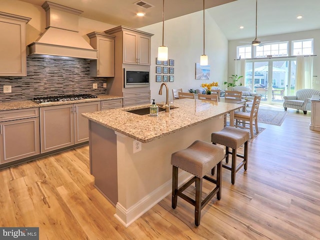 kitchen featuring a center island with sink, custom range hood, decorative light fixtures, stainless steel gas stovetop, and a kitchen bar