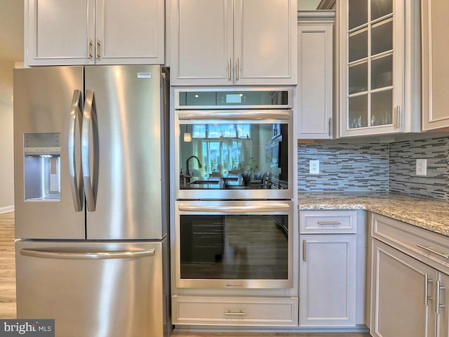 kitchen featuring appliances with stainless steel finishes, glass insert cabinets, light stone counters, and tasteful backsplash