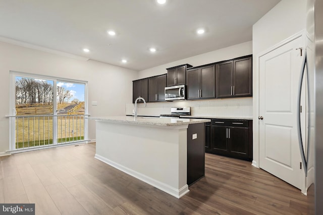 kitchen featuring stainless steel appliances, a sink, light stone countertops, dark wood-style floors, and an island with sink