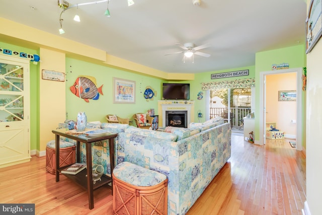 living area featuring light wood-type flooring, a glass covered fireplace, ceiling fan, and baseboards