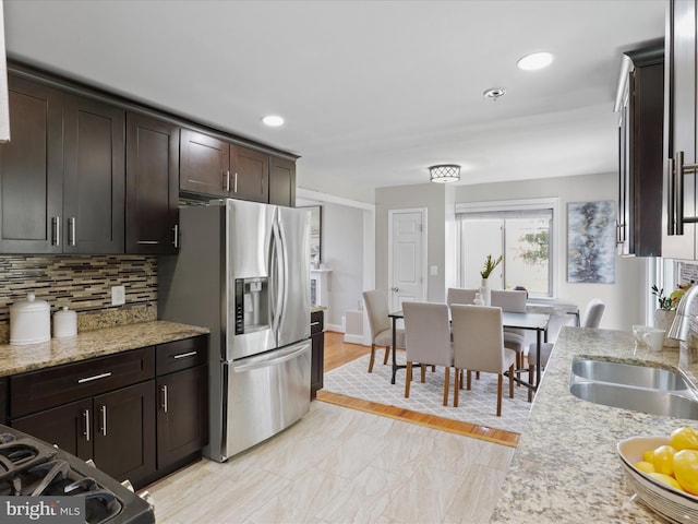 kitchen with backsplash, light stone countertops, dark brown cabinetry, stainless steel fridge, and a sink