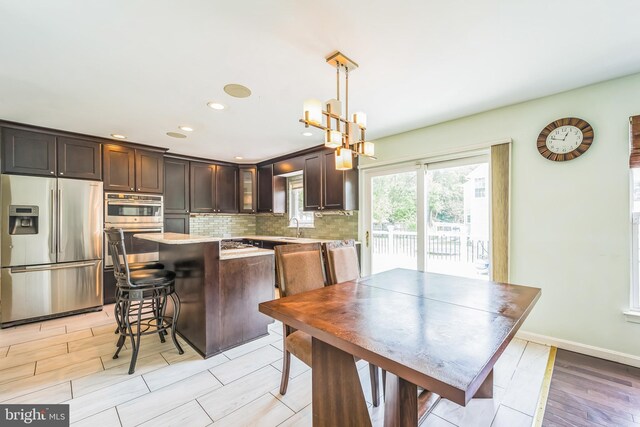 kitchen with light wood-type flooring, appliances with stainless steel finishes, dark brown cabinets, a kitchen island, and tasteful backsplash