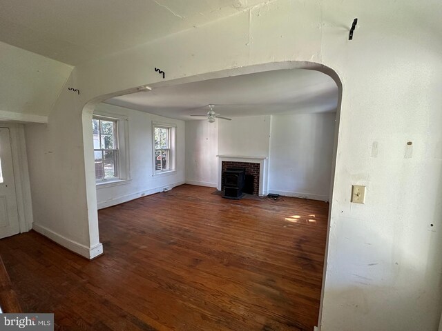 unfurnished living room with arched walkways, dark wood-style flooring, a wood stove, ceiling fan, and baseboards