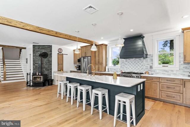 kitchen featuring custom exhaust hood, a kitchen island with sink, light hardwood / wood-style floors, and a wood stove