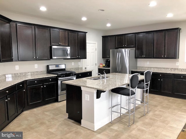 kitchen featuring a center island with sink, sink, light stone countertops, appliances with stainless steel finishes, and a kitchen bar