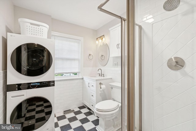 bathroom with a shower, stacked washing maching and dryer, tile walls, vanity, and toilet