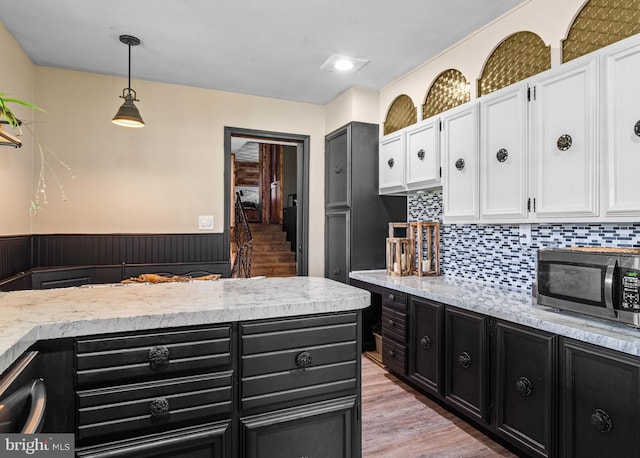 kitchen featuring light wood-type flooring, stainless steel microwave, dark cabinetry, white cabinetry, and hanging light fixtures