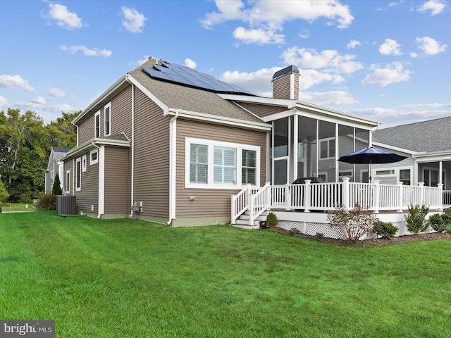 rear view of property featuring a wooden deck, central AC unit, a sunroom, solar panels, and a yard