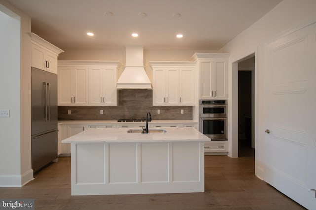 kitchen with dark wood-type flooring, an island with sink, backsplash, stainless steel appliances, and premium range hood