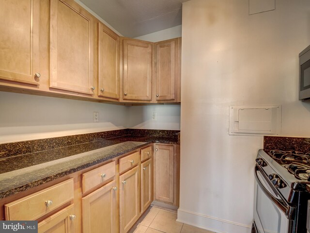 kitchen with light brown cabinetry, dark stone countertops, light tile patterned floors, and white range with gas cooktop