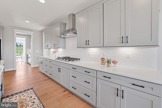 kitchen with light wood finished floors, decorative backsplash, light countertops, wall chimney range hood, and stainless steel gas stovetop