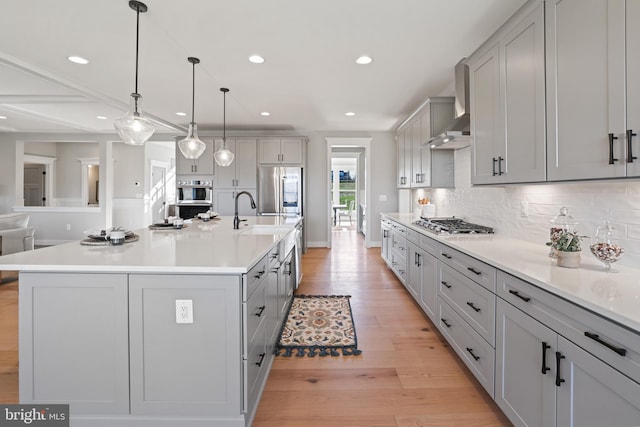 kitchen with gray cabinetry, stainless steel appliances, wall chimney range hood, decorative backsplash, and light wood finished floors